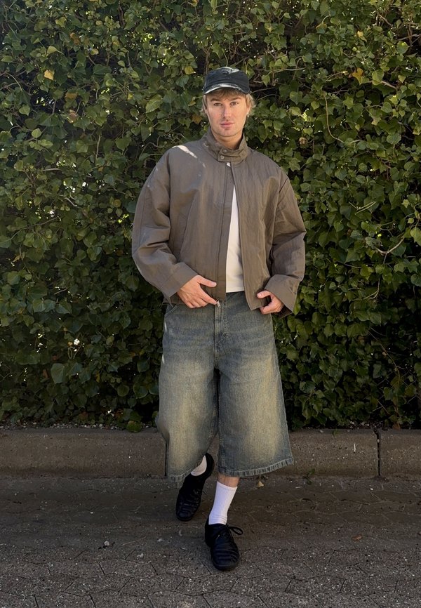 Jeune personne portant une veste courte marron, un short en jean large, une casquette noire, des chaussettes blanches et des chaussures noires, debout à l'extérieur devant un fond feuillu.