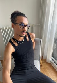 Débardeur noir à côtes avec épaules découpées, associé à un pantalon noir. Les accessoires comprennent un collier en chaîne et des lunettes rondes. Fond clair.