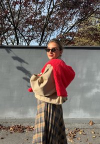 Beige oversized jas gecombineerd met een rood gebreide trui en een geruite maxi-rok. De scène toont een grijze muur en gevallen bladeren.