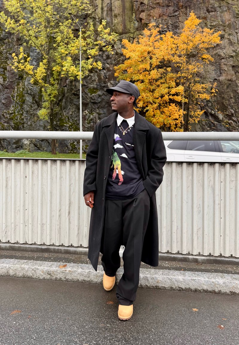 Manteau noir par-dessus un sweat-shirt imprimé, pantalon noir ample et sabots beige. La scène présente un feuillage d'automne et des éléments rocheux en arrière-plan.