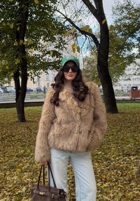 Lažna krzna jakna u svijetlosmeđoj boji, s luksuznom teksturom i oversized krojem, uparena s plavim farmerkama i tamnosmeđom torbom.