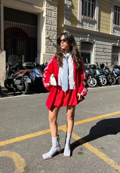 Chaqueta atlética roja con rayas blancas, camisa blanca, corbata de rayas azules, falda roja y botas de tobillo plateadas, con un fondo de calle.