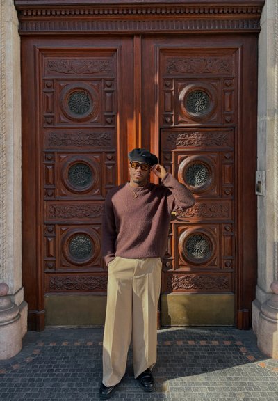 Suéter marrón de punto, pantalones de pierna ancha color crema, boina negra y gafas de sol. De pie frente a unas puertas dobles de madera detalladas con motivos circulares.