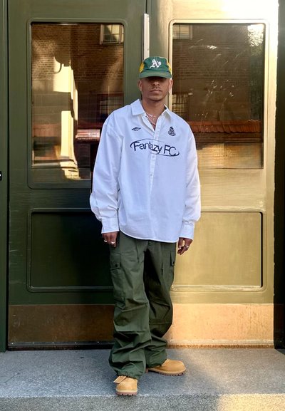 Camisa blanca de manga larga con el logo de "Fantasy FC", pantalones cargo verdes, botas beige y una gorra verde y amarilla. De pie frente a puertas de cristal.