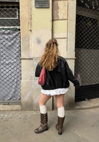 Giacca oversize in pelle nera, vestito bianco con ruches, stivali marroni al ginocchio con fibbie e una piccola borsa bordeaux. Sfondo urbano.