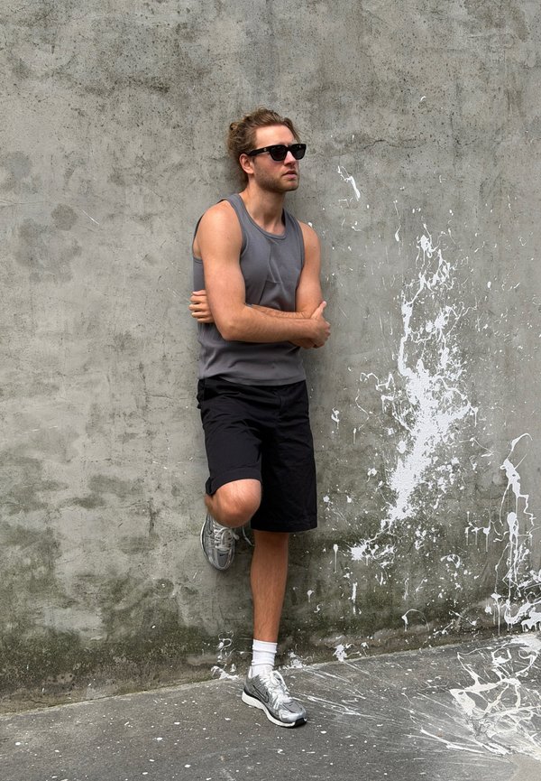 Man met zonnebril, grijs tanktopje, zwarte shorts en sneakers, leunt tegen een betonnen muur met witte verfvlekken.