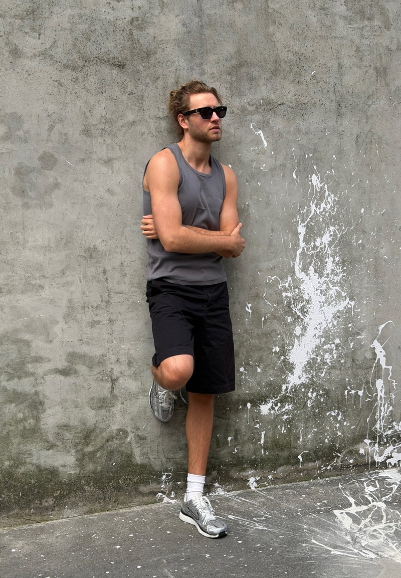Man som bär solglasögon, grå tanktopp, svarta shorts och sneakers lutar sig mot en betongvägg med vita färgstänk.