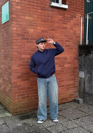 Joven con chaqueta azul marino, vaqueros anchos y gorra a rayas, apoyado contra una pared de ladrillos rojos cerca de un cartel que dice "Área de Montaje A".