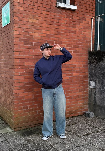 Joven con chaqueta azul marino, vaqueros anchos y gorra a rayas, apoyado contra una pared de ladrillos rojos cerca de un cartel que dice "Área de Montaje A".
