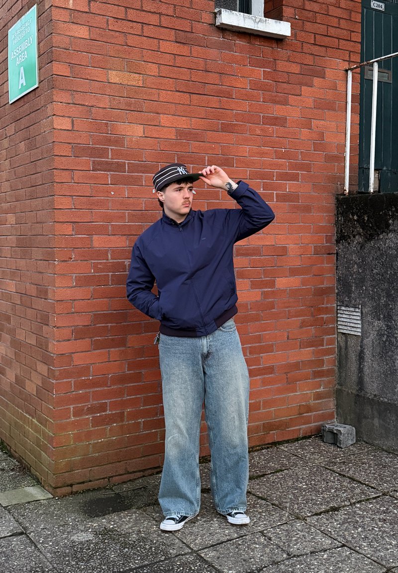 Joven con chaqueta azul marino, vaqueros anchos y gorra a rayas, apoyado contra una pared de ladrillos rojos cerca de un cartel que dice "Área de Montaje A".