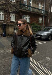 Femme portant une veste en cuir noire avec bordure en fourrure, lunettes de soleil et jean, debout dans une rue de la ville avec une voiture garée et un bâtiment recouvert de lierre en arrière-plan.