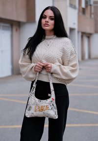 Maglione beige lavorato a maglia con motivo a treccia, pantaloni neri a vita alta e una borsa bianca con grafiche e "DESIGNS" in oro.