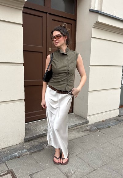 Mujer con gafas de sol, top sin mangas color oliva, falda blanca de satén, cinturón marrón, chanclas, y lleva un bolso de hombro negro, de pie en la acera.
