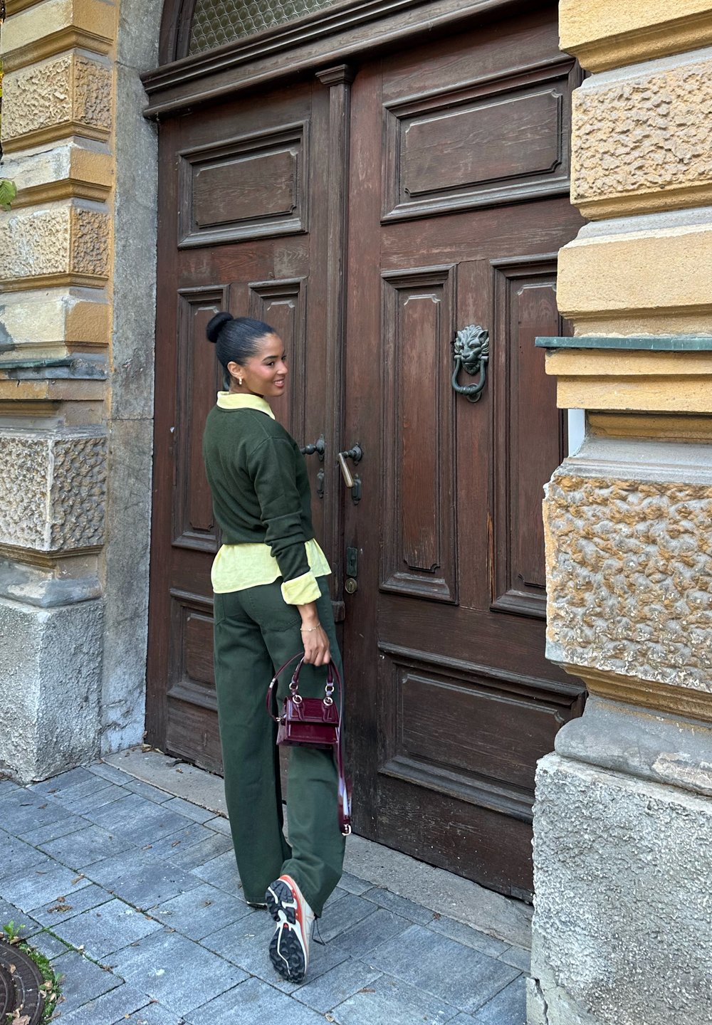 Green velour sweater and matching trousers layered over a yellow shirt, holding a small burgundy handbag, with a wooden door in the background.