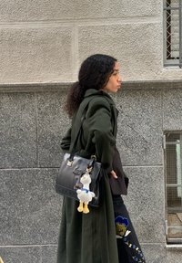 Melna ādas rokassoma ar noņemamu mīksta rotaļlieta piekariņu, kuru nes cilvēks garā olīvzaļā mētelī, ar sarežģītiem zilo un dzelteno izšuvumu motīviem uz biksēm.