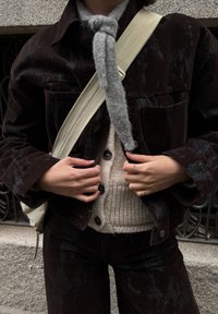 Tamsiai ruda kordurovo striukė su tekstūruotu paviršiumi, derinama su šviesiai smėlio spalvos megztiniu. Pilkas šalikas ir smėlio spalvos dirželis per petį.