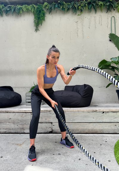 Una mujer con un sujetador deportivo lavanda y pantalones deportivos negros agarra una gruesa cuerda de batalla retorcida sobre una superficie de concreto, con sacos de frijoles negros cerca.