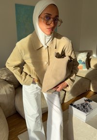 Casaco de camurça cor de castanho claro com fecho de botões e calças largas brancas. Segura uma mala de tecido bege, usa óculos grandes e um hijab branco.