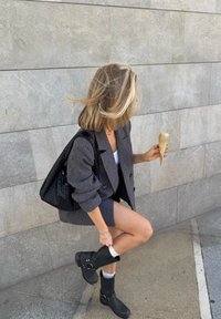 Blazer gris sobre una camiseta blanca, combinado con una falda corta gris. Bolsa de mano tejida negra y botas negras hasta la mitad de la pantorrilla. Sostiene un cucurucho con helado de color claro.