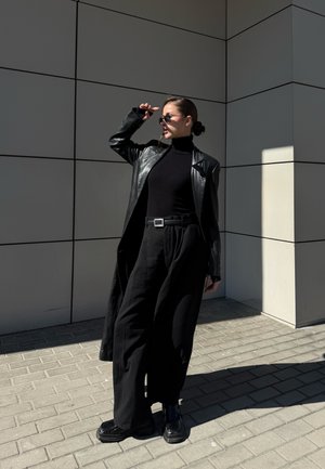 Mujer con pantalones negros de pernera ancha, cuello alto y abrigo de cuero ajusta sus gafas de sol mientras está de pie junto a un edificio moderno con azulejos bajo la luz del sol.