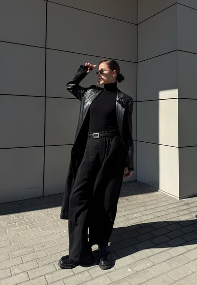 Mujer con pantalones negros de pernera ancha, cuello alto y abrigo de cuero ajusta sus gafas de sol mientras está de pie junto a un edificio moderno con azulejos bajo la luz del sol.