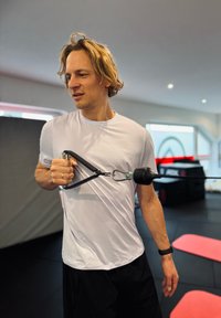 Fehér pólós férfi fekete markolattal ellátott fitnesz kábelgép használatával. Az edzőterem fekete gumiszőnyeggel és piros edzőszőnyegekkel rendelkezik.