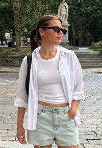 Λευκό πουκάμισο με κουμπιά, στρωμένο πάνω από ένα λευκό μπλουζάκι με ρίγες, συνδυασμένο με ανοιχτόχρωμο τζιν σορτς. Τα αξεσουάρ περιλαμβάνουν γυαλιά ηλίου και μία χρυσή αλυσίδα. 
