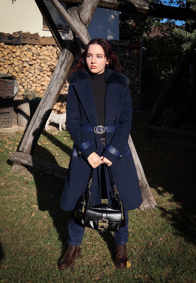 Cappotto di lana navy con collo in pelliccia sintetica, cintura arricciata e dettagli in oro. Abbinato a jeans scuri, stivali marroni e una borsa in pelle nera.
