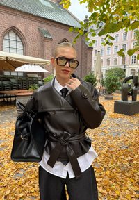 Fekete bőr trench coat, dupla gombolású dizájnnal, túlméretezett gombokkal, övvel a deréknál, fehér ing és fekete nyakkendő fölött viselve. Fekete táska a kézben.