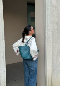 Tkaninska bistra torba s dvostrukim ručkama, s teksturiranim završetkom. Bijela košulja dugih rukava s crvenim i plavim vezenim detaljima, plave traperice.
