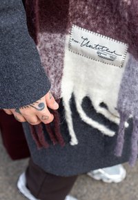 Ruudullinen villakangastakki tummanharmaana ja viininpunaisena, pehmeällä pintakäsittelyllä, hapsureunuksin ja valkoisella käsin kirjoitetulla tekstillä varustetulla nimilapulla.