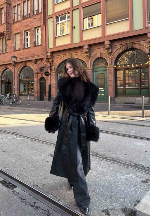 Ung kvinde i sort læderjakke med pelskant, der går på sporvognsskinner foran historiske europæiske bygninger med buede vinduer og cykler.