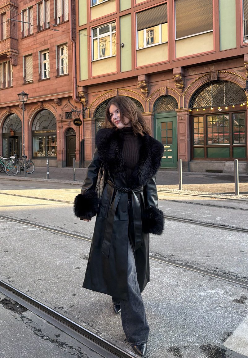 Jeune femme en manteau en cuir noir avec bordure en fourrure marchant sur des voies de tram devant des bâtiments historiques européens aux fenêtres cintrées et des bicyclettes.