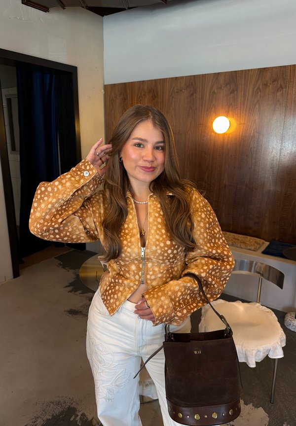 Une femme pose dans une veste brune à motifs et un pantalon blanc, tenant un sac seau foncé dans une pièce lambrissée.