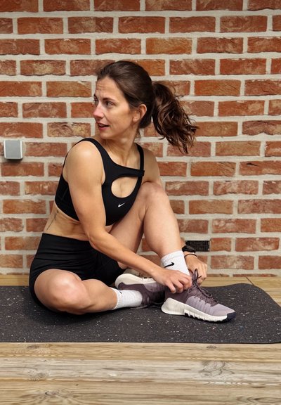 Mujer con ropa deportiva negra atándose los cordones de un pie, sentada sobre una esterilla de ejercicio contra una pared de ladrillos.