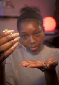 Genomskinlig pipett hålls ovanför en öppen handflata med långa naglar. Bakgrunden har ett mjukt glödande runt ljus och suddiga hyllobjekt.