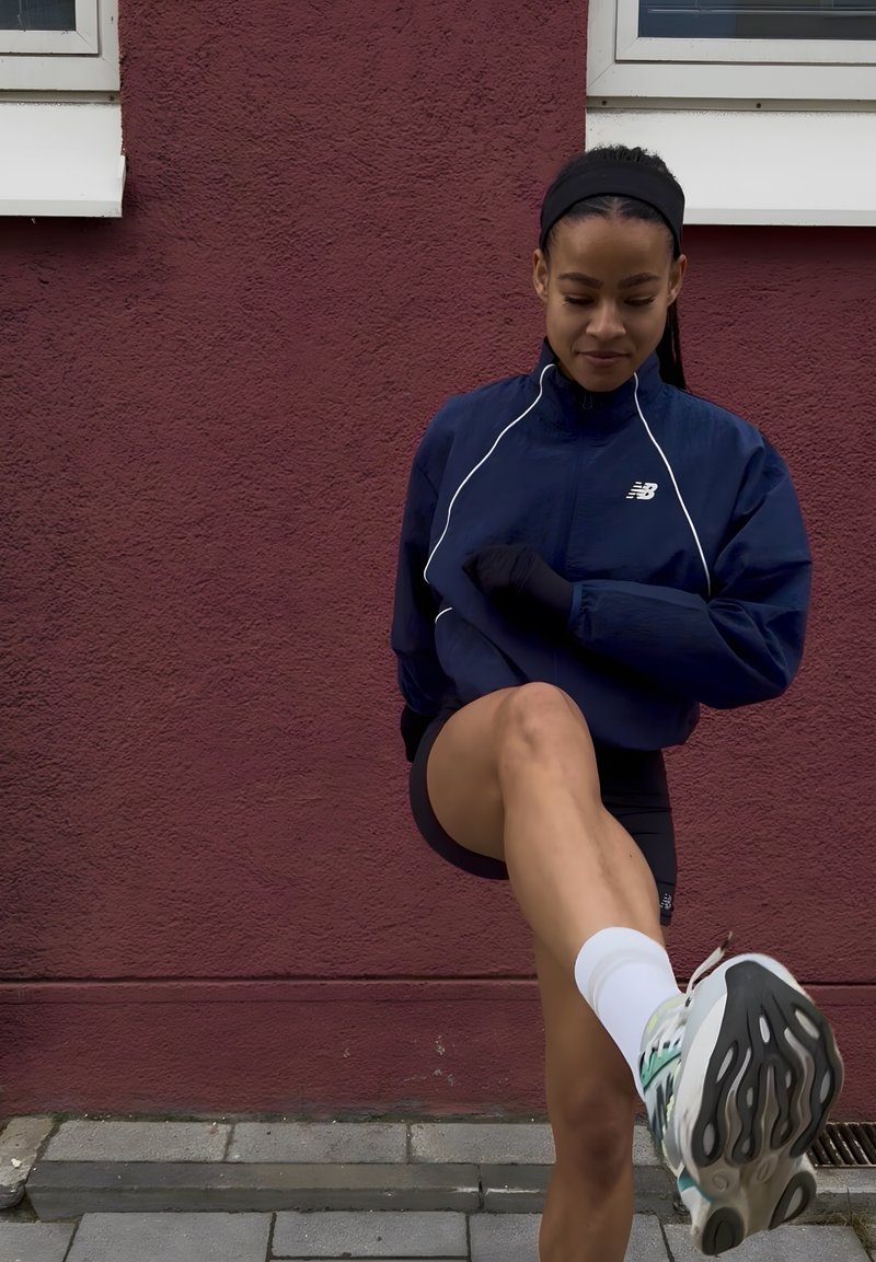 Νεαρή γυναίκα με μπλε μπουφάν και μαύρα σορτς τεντώνει το ένα πόδι μπροστά σε πεζοδρόμιο μπροστά από κόκκινο τοίχο.