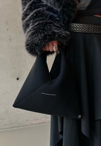 Personne portant un manteau en fourrure foncé et une jupe noire, tenant un sac à main triangulaire noir avec un détail de couture en ligne et un anneau décoratif à la main.
