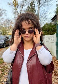 Vīna krāsas ādas veste ar rāvējslēdzēju priekšā, zem tās valkāta balta ar garām piedurknēm krekls, un kaķa acu formas saulesbrilles. Uz rokas brūns teksturēts viedpulkstenis.