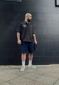Črna majica s kratkimi rokavi in belim ovratnikom ter logotipom, modri denim kratke hlače, bele nogavice, šahovske slip-on čevlje, drži črno kapo.