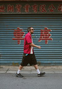 Camisa roja de manga corta con detalles blancos, pantalones cortos negros, calcetines blancos y zapatillas negras. Camina frente a una persiana azul con texto rojo.