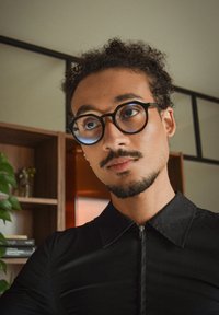 Zwart shirt met ritssluiting en kraag, gedragen door een persoon met krullend haar en een bril, voor een achtergrond van houten planken en planten.