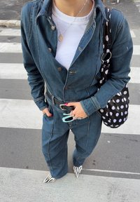 Άτομο με ολόσωμη φόρμα από τζιν και λευκό πουκάμισο που κρατά ένα μπλε κλιπ, φορά παπούτσια με ριγέ ζέβρας και έχει μια μαύρη-λευκή τσάντα με πουά.