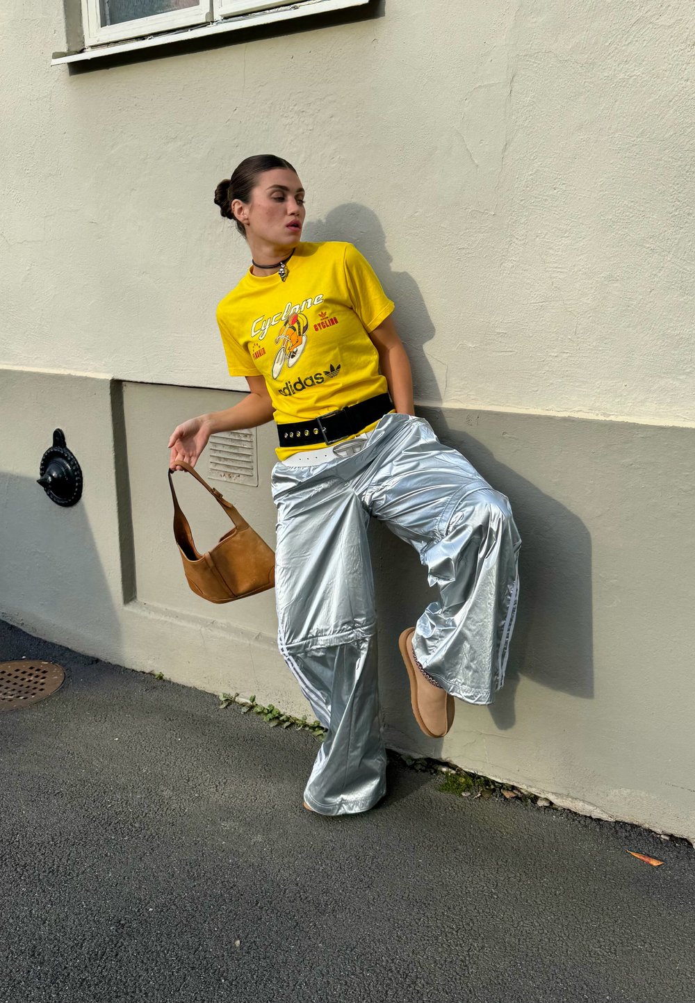 Žlté grafické tričko s nápisom "Cyklón", strieborné voľné nohavice, čierny opasok a hnedá kabelka cez rameno. Dokončené neutrálnej farby topánkami a náhrdelníkom.