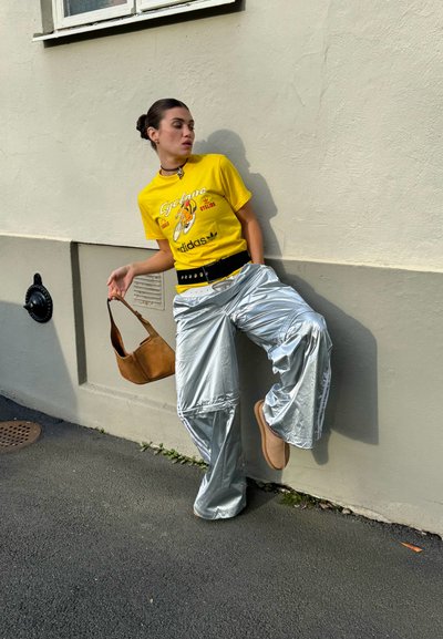 Camiseta gráfica amarilla con estampado "Cyclone", pantalones plateados holgados, cinturón negro y bolso marrón de hombro. Combinado con zapatos neutros y choker.