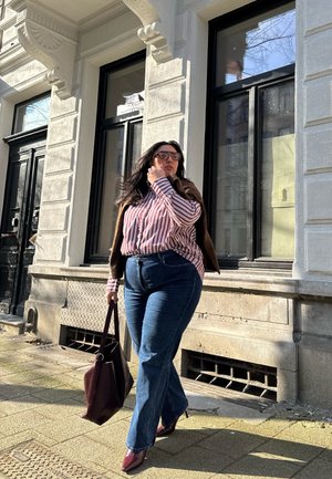 Una persona con una camiseta de rayas rojas y blancas, un cárdigan marrón, unos vaqueros acampanados azules y tacones burdeos, llevando un bolso texturizado burdeos, caminando al aire libre.