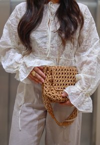 Femme en blouse blanche brodée tenant un sac à main en perles de bois beige avec une bague assortie, portant un pantalon de couleur claire.