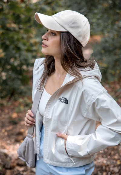 Chaqueta ligera blanca con capucha y logo en el lado izquierdo del pecho, combinada con una gorra beige y un bolso gris de hombro, todo en un entorno de vegetación.