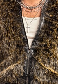 Μαύρο δερμάτινο τζάκετ με καφέ γούνα στον γιακά, φερμουάρ στο μπροστινό μέρος, πο�λλαπλές ασημένιες αλυσίδες και κόσμημα με σταυρό πάνω από ένα λευκό φανελάκι.