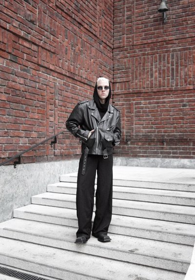Persona con cabello rubio corto que lleva una chaqueta de cuero negra, pantalones anchos negros, gafas de sol y capucha, de pie en unas escaleras de concreto junto a una pared de ladrillo rojo.